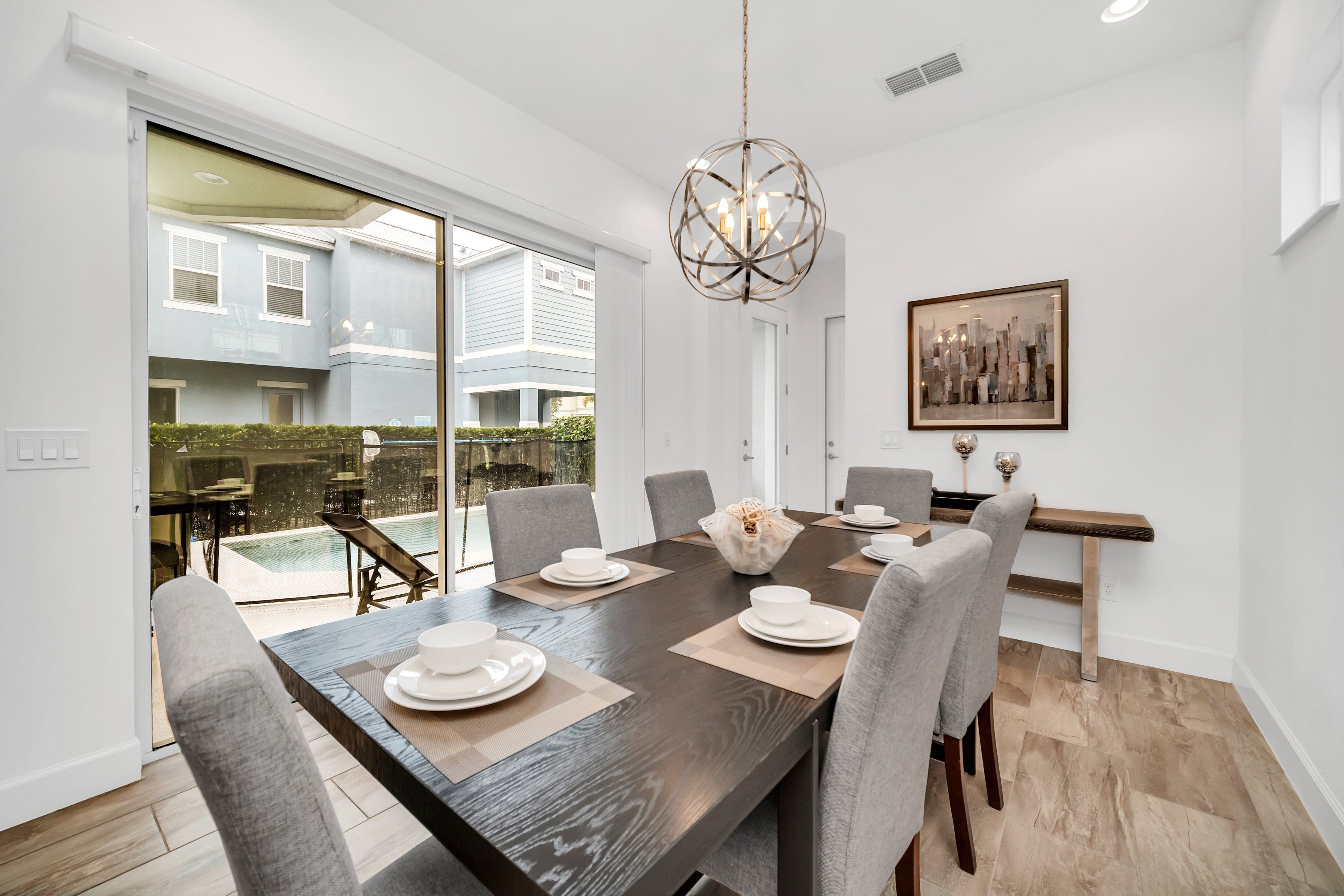 Comedor con una mesa de madera oscura dispuesta para seis personas, rodeada de sillas grises. Un ventanal ofrece vistas a una piscina y una casa azul.
