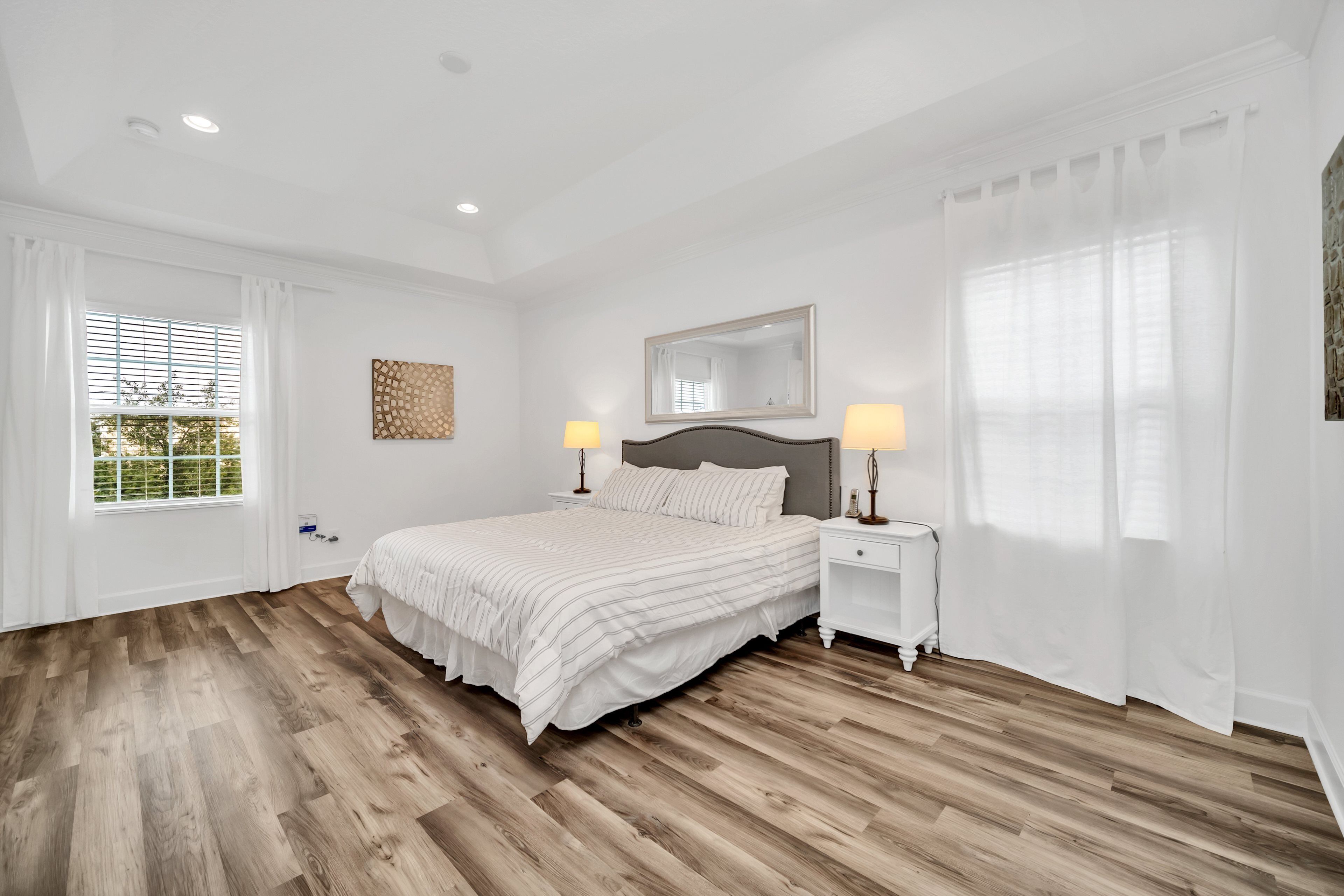 Une chambre à coucher avec un grand lit, deux tables de chevet avec des lampes, une tête de lit, une fenêtre avec des stores et un miroir fixé au mur.