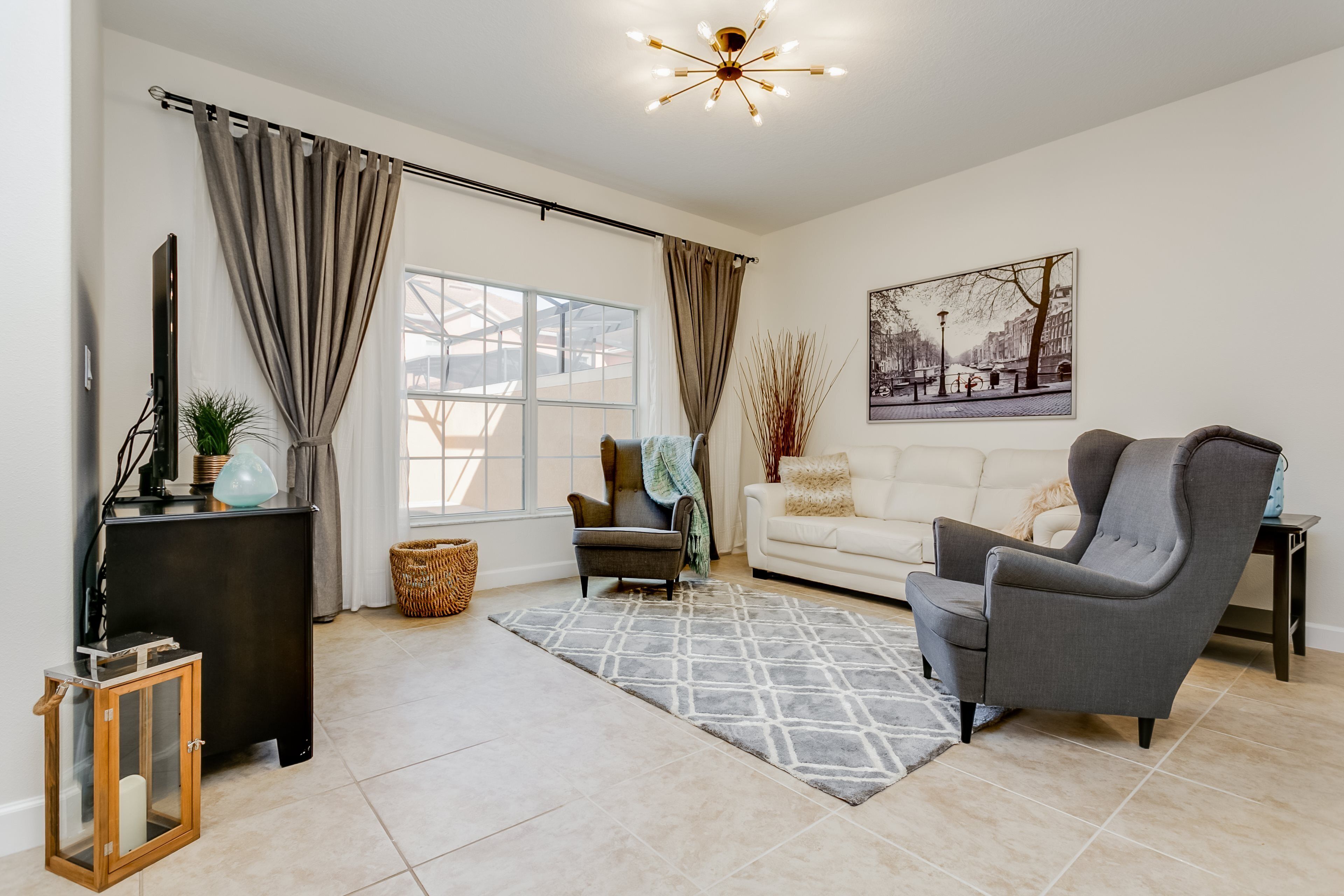 A modern living room with a grey armchair, a white sofa, a large window with curtains, a floor lamp, and a chandelier.