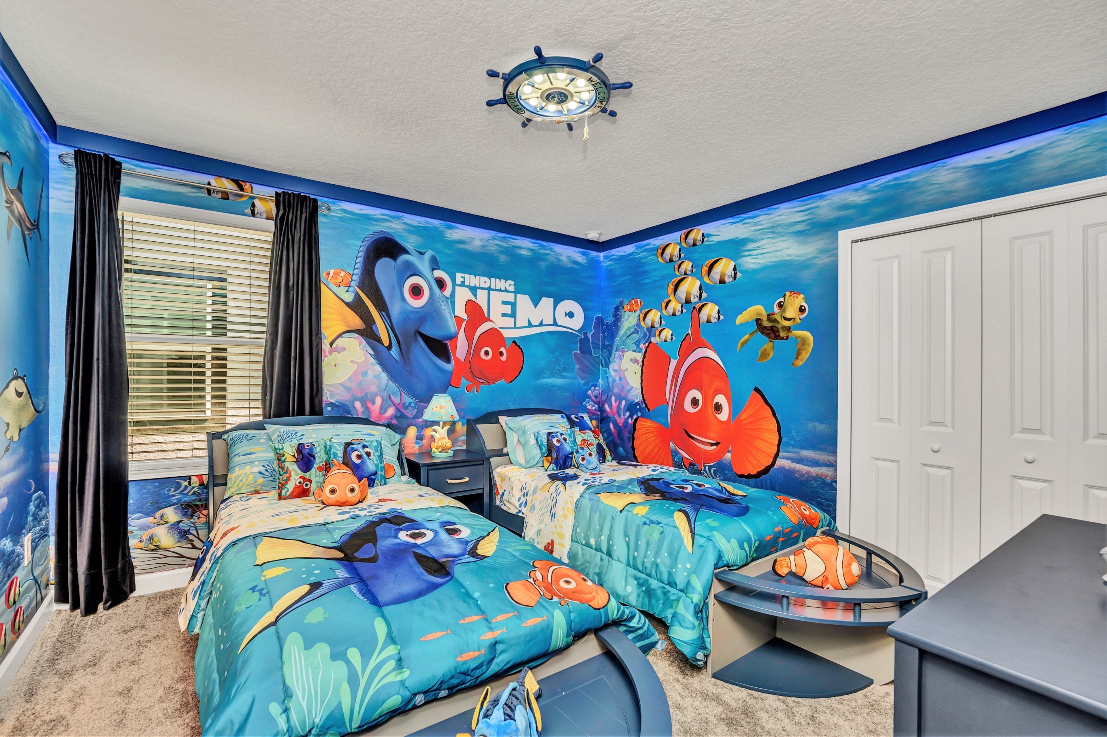 Ein Zimmer mit zwei Betten, einem Schreibtisch und einem Deckenventilator. Die Wände sind mit einem Wandbild aus Findet Nemo verziert.