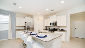 A modern kitchen with white cabinets, a stainless steel refrigerator, a microwave, and a gas stove. There are four white bar stools around the island. - 5 Bdrm Solterra New Home (Davenport)