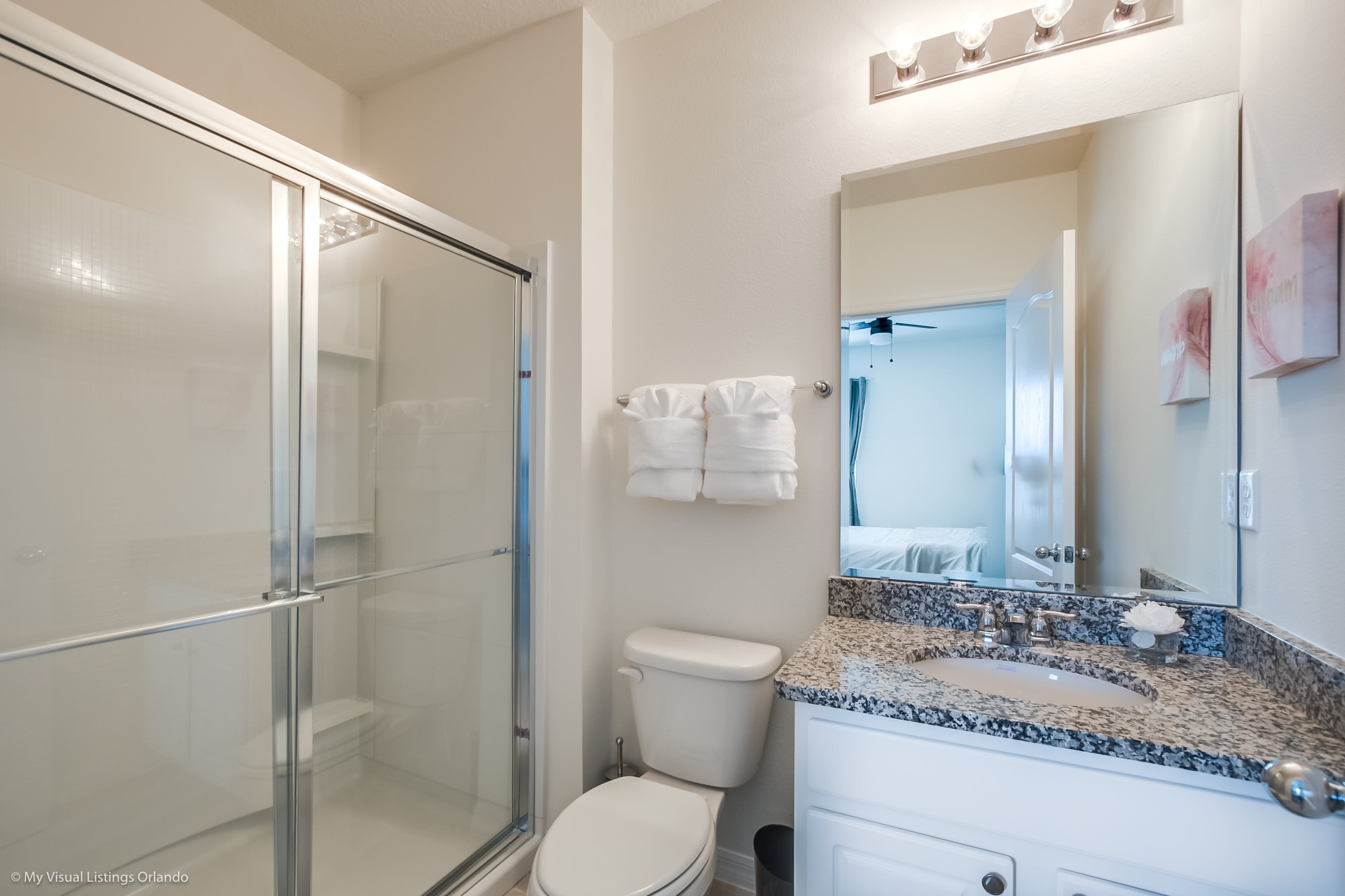 Un baño moderno con cabina de ducha de vidrio, inodoro blanco, lavamanos sobre encimera de granito y un espejo grande.