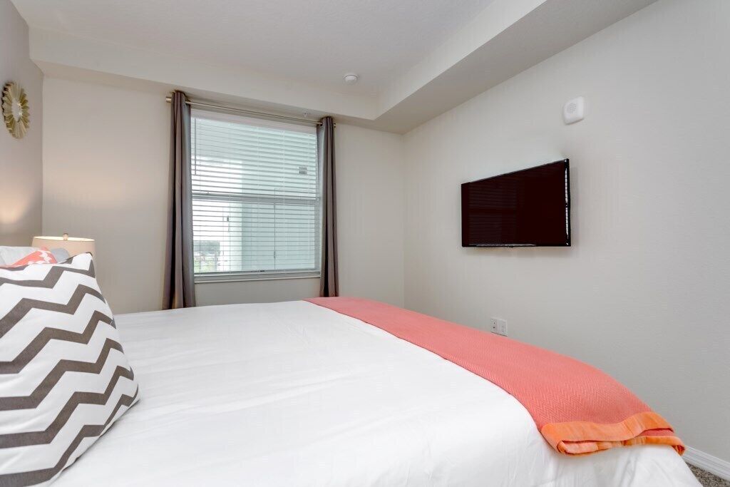 Una habitación de hotel con cama, televisor montado en la pared y ventana con cortinas.