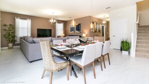 A modern living room with a sofa, dining table, and chairs. There is a flat-screen TV mounted on the wall, a dining table set with plates and glasses, and a staircase leading to another level. - 6 Bedroom, 6 Bath Pool Home (Davenport)