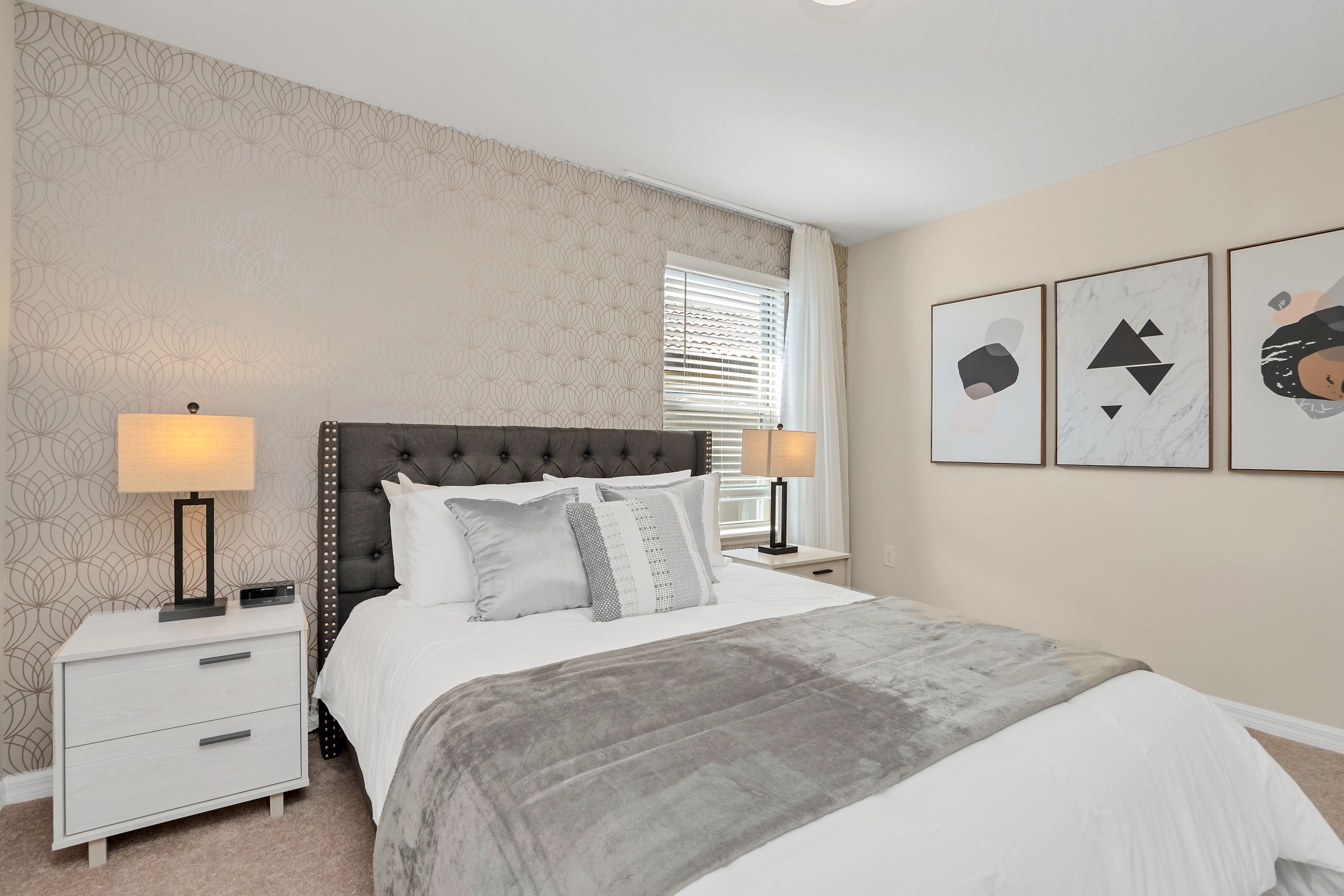 A bedroom with a tufted headboard, a bed with white bedding, two bedside tables with lamps, a window with blinds, and three framed artworks on the wall.