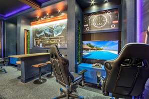 A modern gaming room with two gaming chairs, a desk, and a large screen displaying a tropical beach scene.