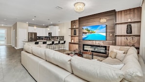 A modern living room with a large flat-screen TV, a beige sectional sofa, a dining area with bar stools, and a kitchen in the background.