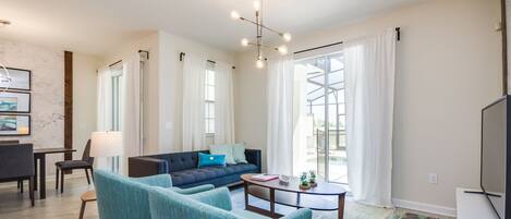 Una sala moderna con un sofá, dos sillones, una mesa de centro y una televisión. Hay un comedor con una mesa y sillas, y ventanas grandes con cortinas.