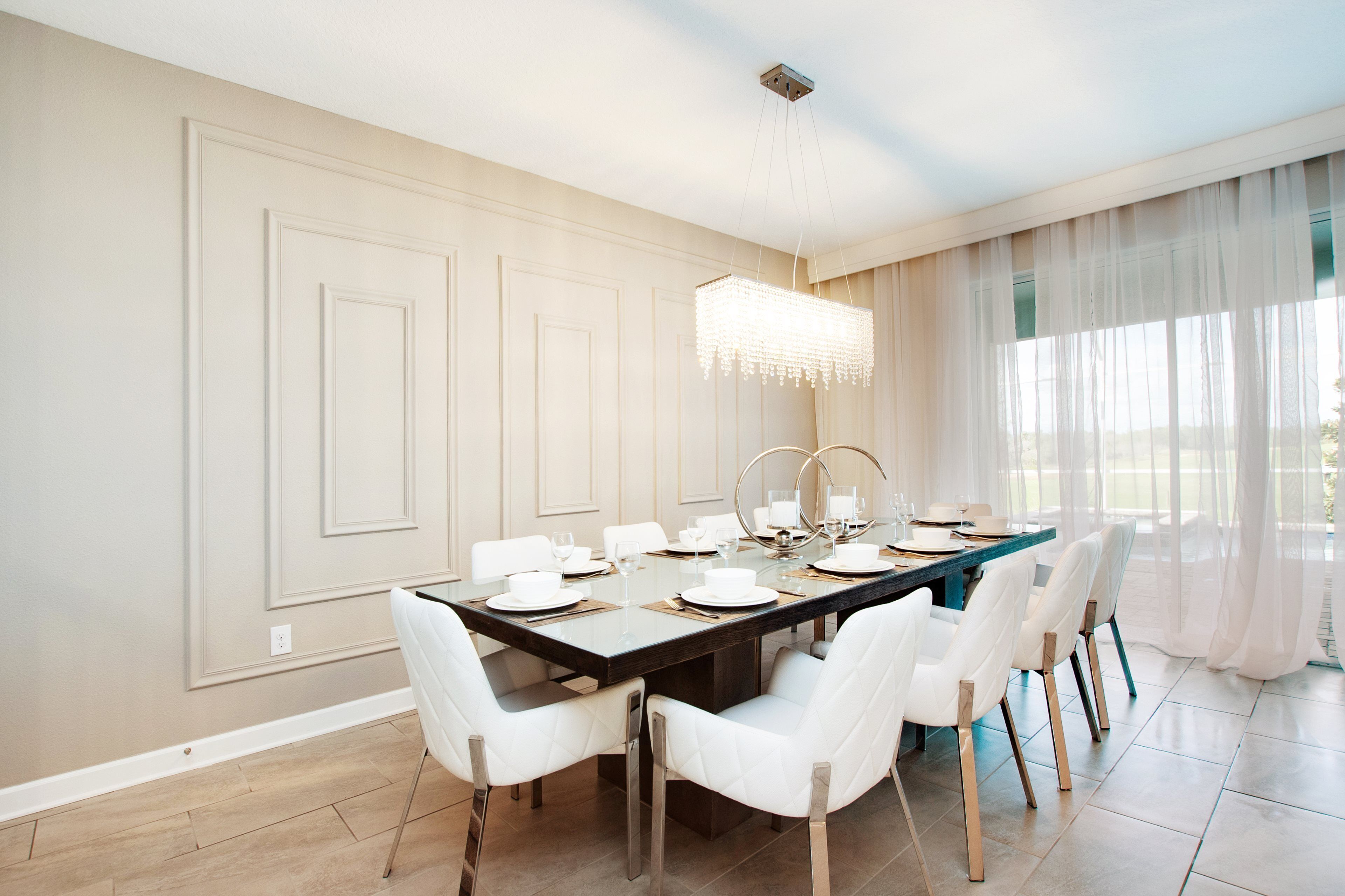 Uma sala de jantar com uma mesa comprida posta para seis pessoas, cadeiras brancas, um candelabro e vista através de cortinas leves.
