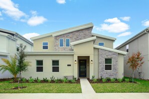 A modern two-story house with a stone facade, a front entrance with a door, and a small garden with various plants. - 9BR Villa with Game Room & Theater (Kissimmee)