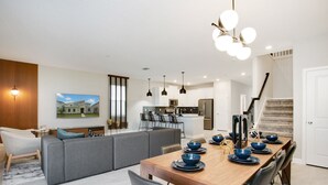 A modern living room with a dining area, a kitchen in the background, and a staircase leading to another floor. - 9BR Villa with Game Room & Theater (Kissimmee)