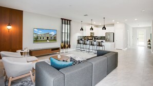 A spacious living area with a grey sectional sofa, a small round coffee table, and a white armchair. A kitchen with modern appliances and bar stools is visible in the background. - 9BR Villa with Game Room & Theater (Kissimmee)