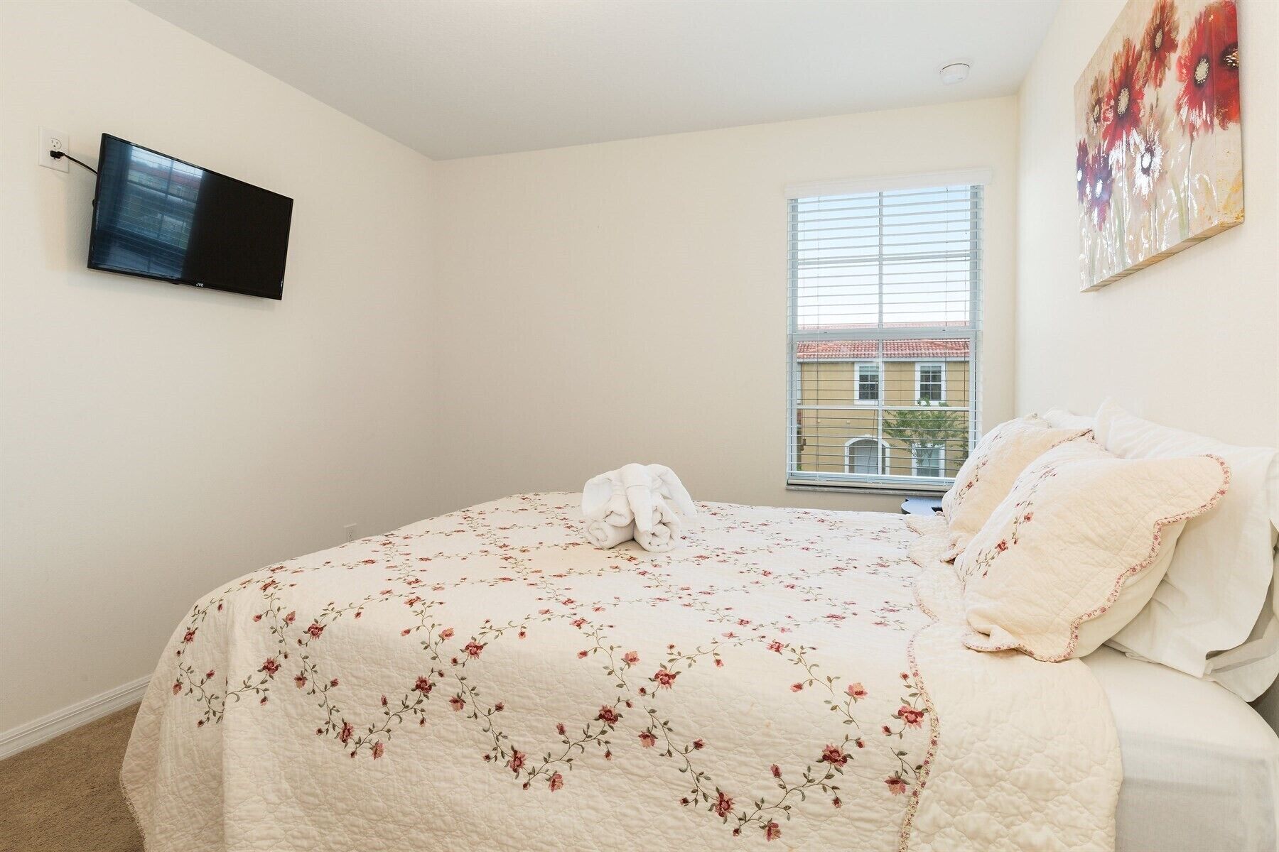 A bedroom with a bed, a flat-screen TV mounted on the wall, a window with blinds, and a painting on the wall.