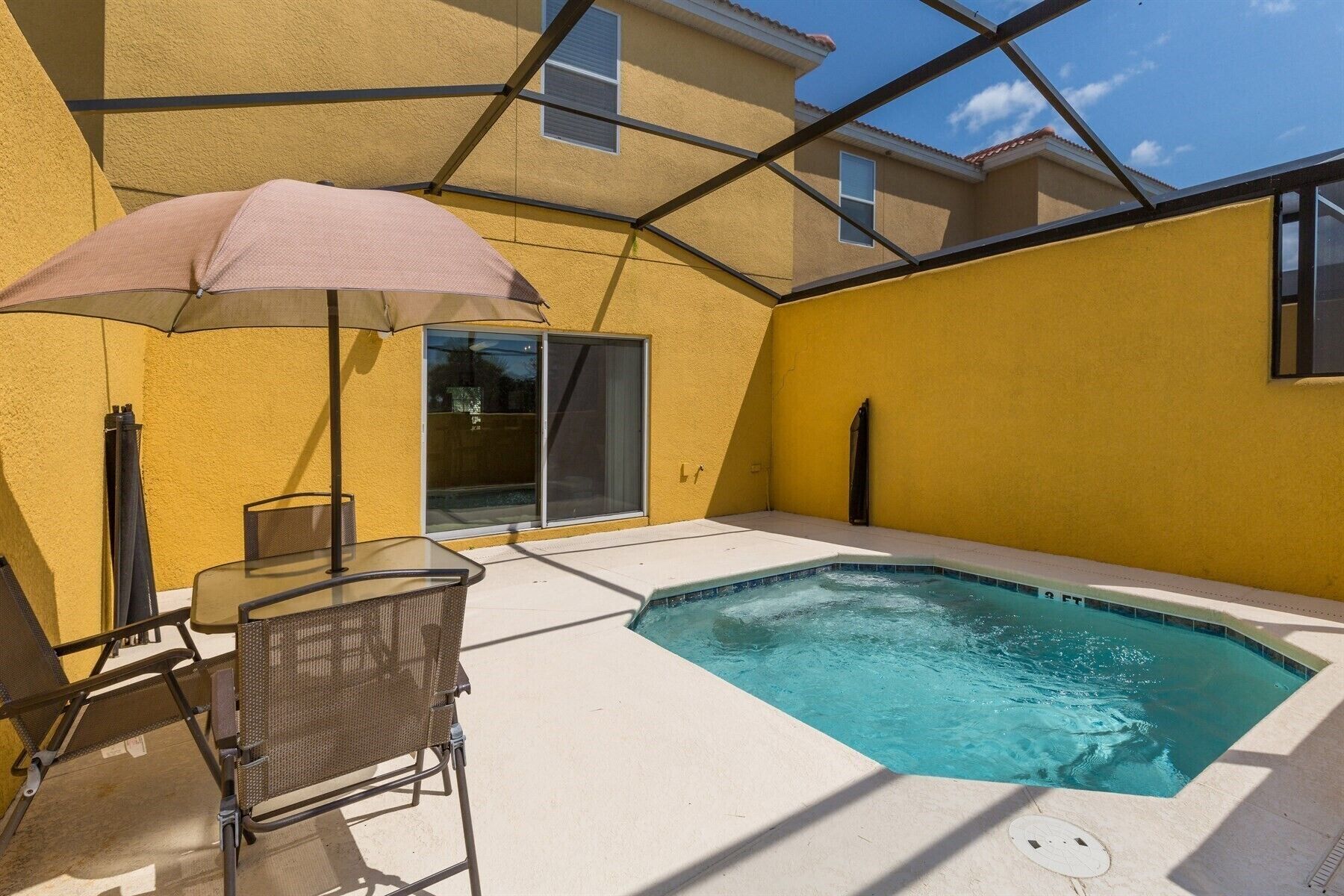 Un petit espace extérieur avec une piscine, une table et des chaises en verre, un parasol et un bâtiment jaune en arrière-plan.