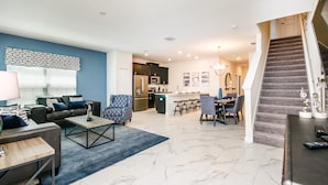 Un salon moderne avec un canapé, une table basse et un coin repas comprenant des chaises et une table. On aperçoit, au fond, une cuisine équipée et un escalier.