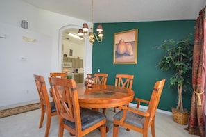 Uma sala de jantar com uma mesa redonda de madeira e seis cadeiras, um lustre, uma parede verde com um quadro emoldurado e um vaso com planta.