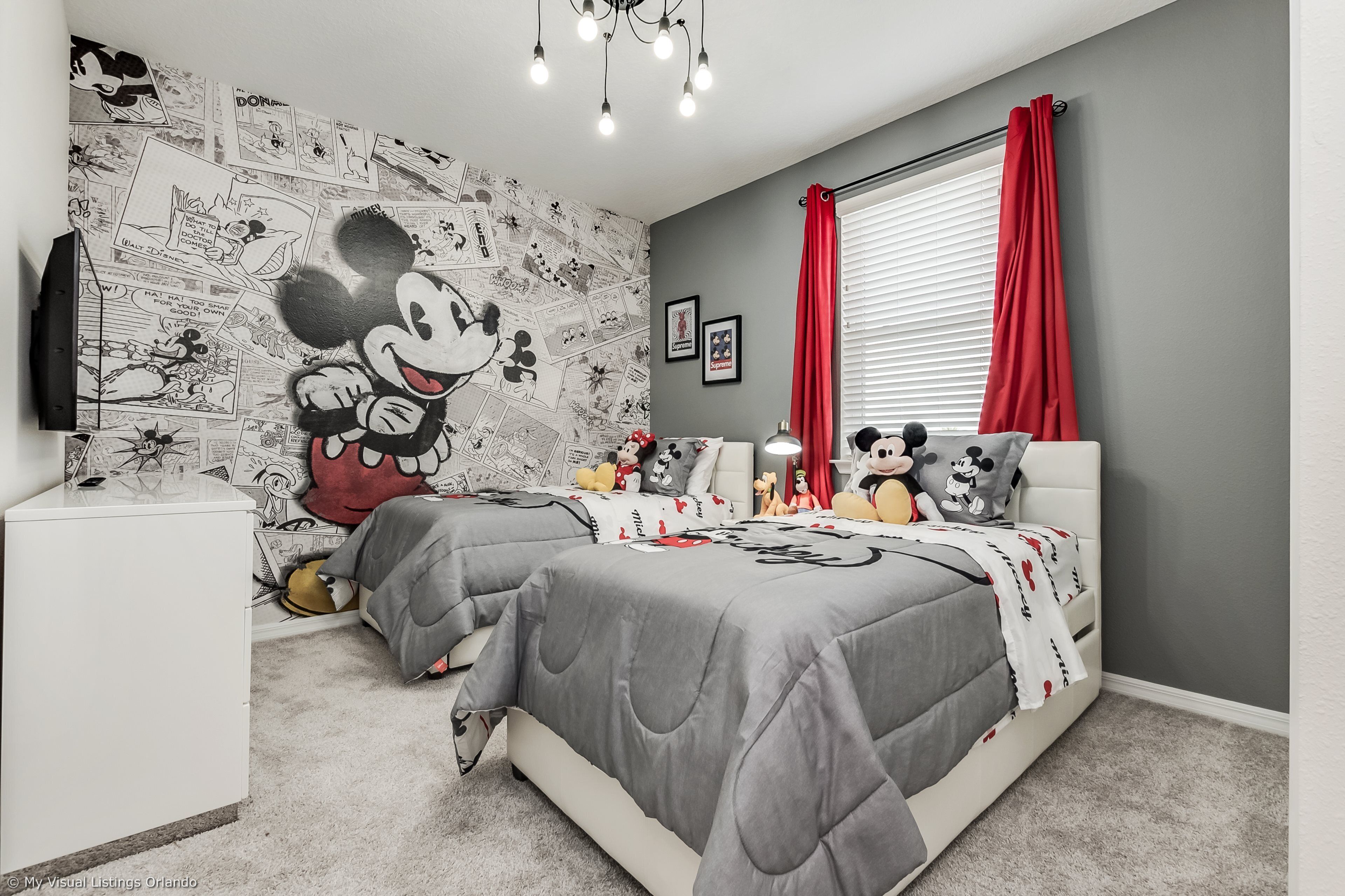 A bedroom with a Mickey Mouse themed wall, a bed with grey bedding, a white bedside table, and a window with red curtains.