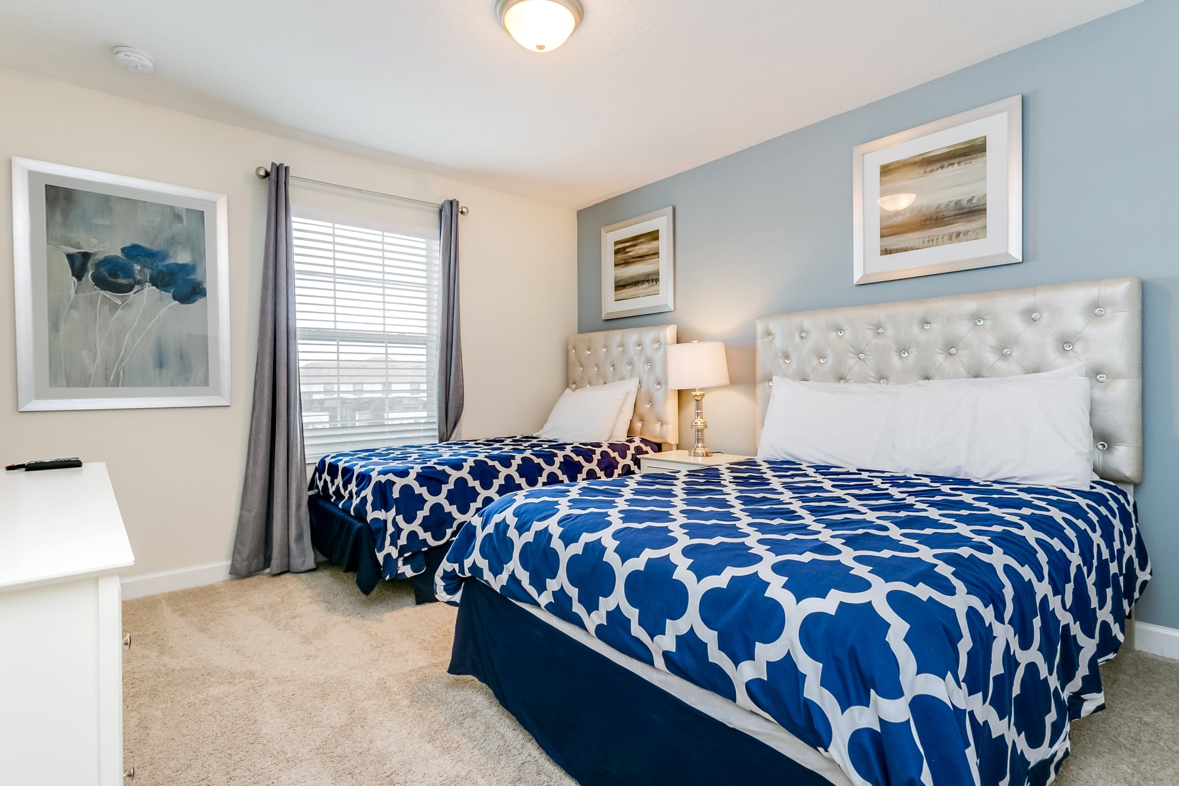 A bedroom with two beds, a bedside table, a lamp, and artwork on the walls.