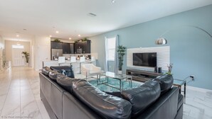 Una sala moderna con un sofá seccional de cuero negro, una mesa de centro de vidrio y un televisor de pantalla plana. Se observa una cocina con gabinetes oscuros y una encimera blanca.