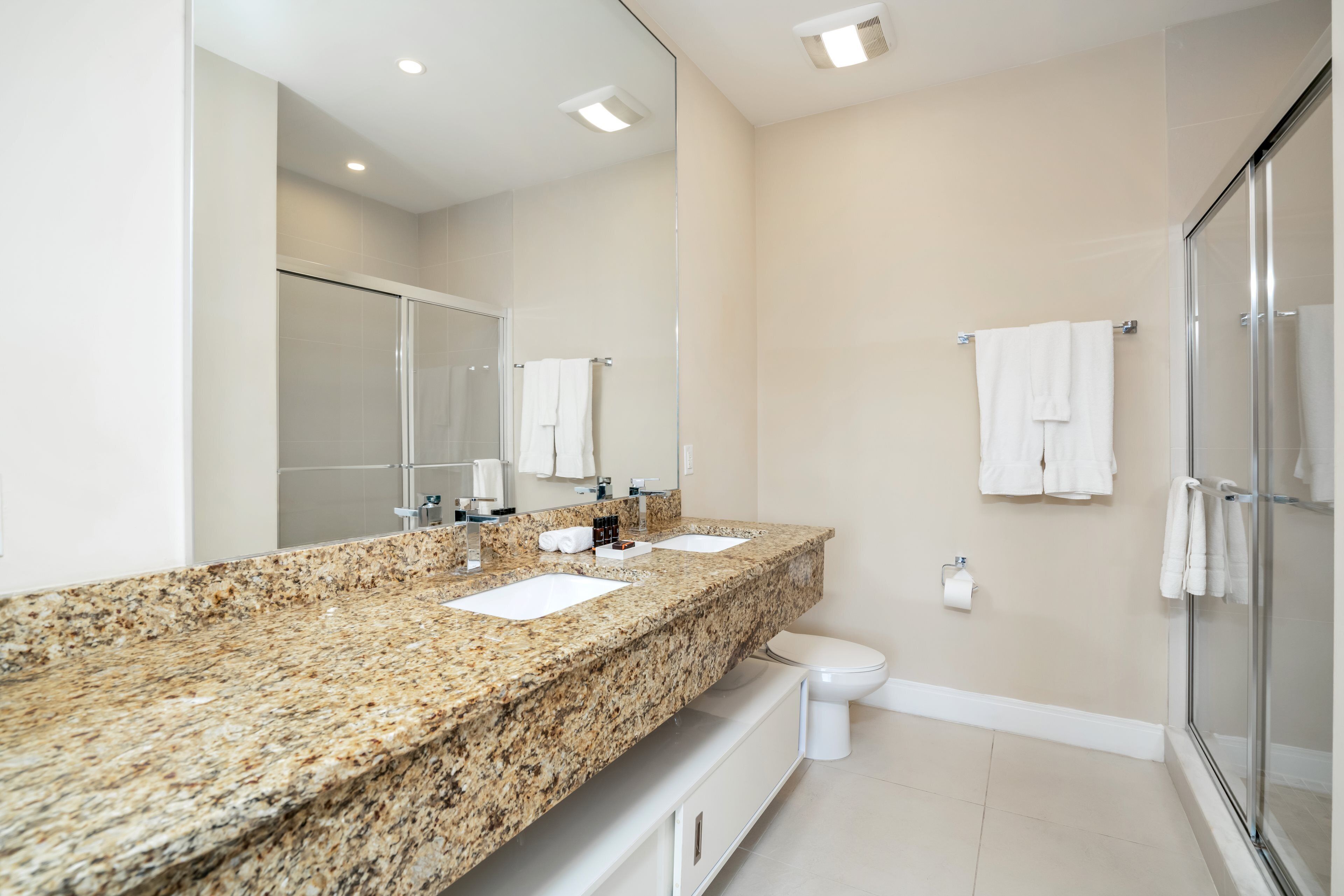 A modern bathroom with a granite countertop, two sinks, a toilet, and a glass shower enclosure.