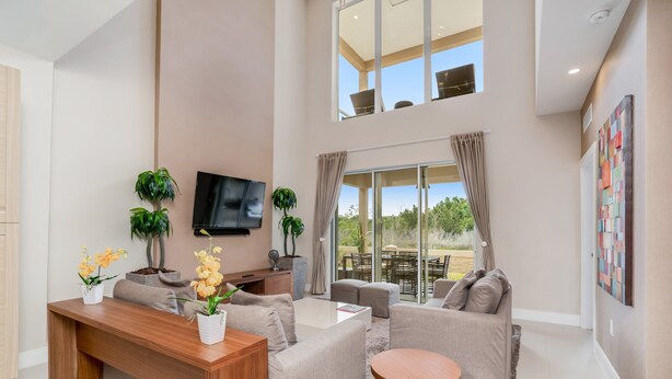 A spacious living area with a wooden desk, beige sofas, and a round table. There are potted plants and a flat-screen TV mounted on the wall.
