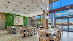 A spacious restaurant with high ceilings, green wall art, and large windows overlooking a pool.