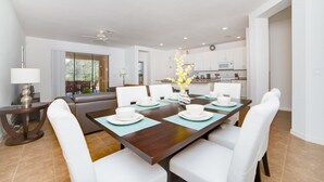 A modern dining area with a dark wooden table set for six, white chairs, and a view of the kitchen in the background. - 2509rose (Davenport)