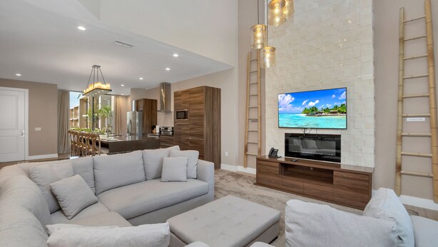 A modern living room with a large sectional sofa, a flat-screen TV mounted on a white brick wall, and a wooden kitchen in the background. - 4bdrm at Magice Village Resort (Kissimmee)