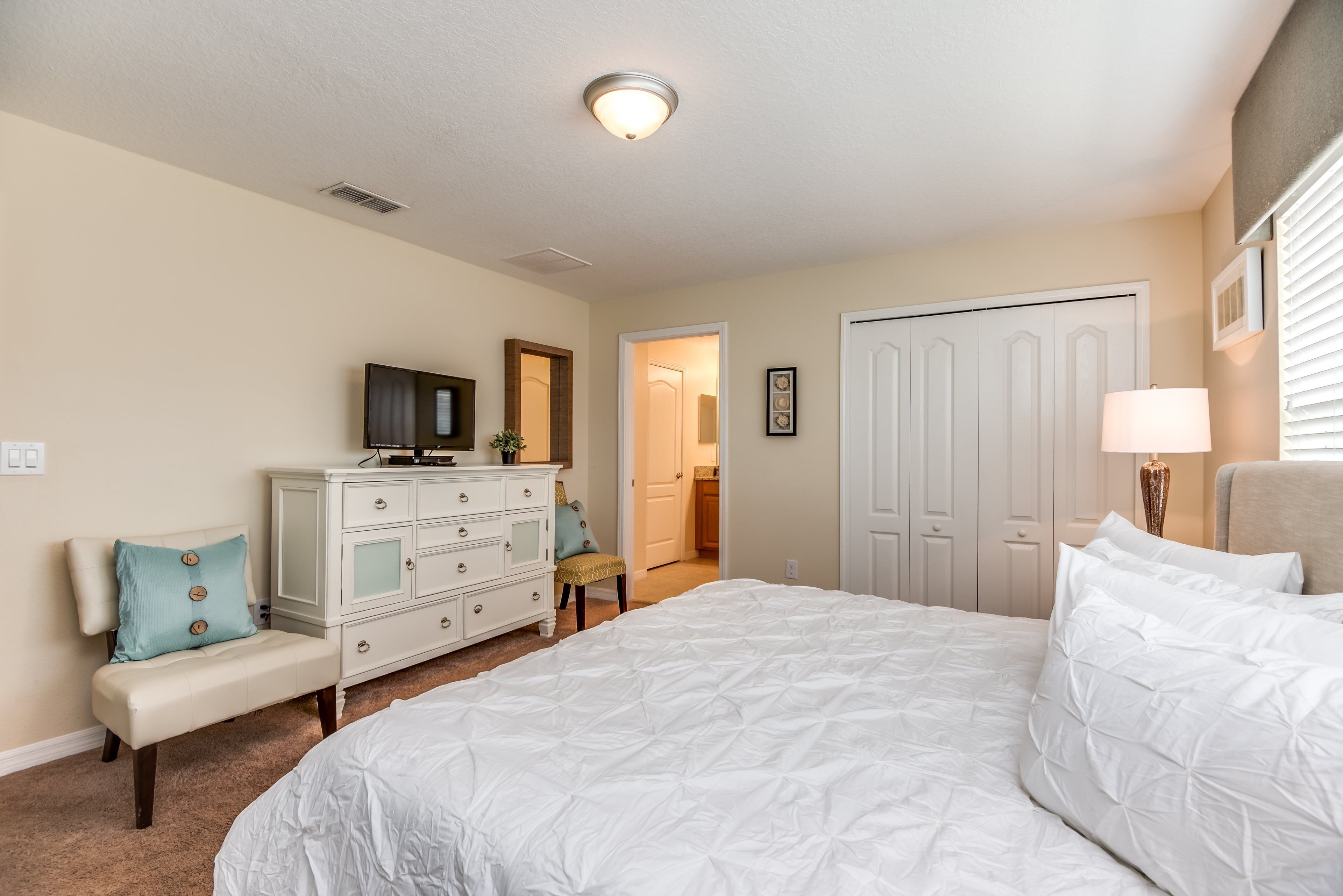 A bedroom with a large bed, a dresser, a chair, and a door leading to another room.