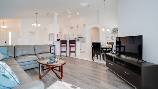 A modern living room with a grey sofa, a coffee table, and a TV on a cabinet. There is a kitchen in the background with white cabinets and a dining area with chairs. - 2360victoria (Davenport)