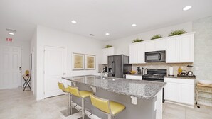 Eine moderne Küche mit weißen Schränken, Granit Arbeitsplatten, einem Edelstahlkühlschrank und einer eingebauten Mikrowelle. Zwei gelbe Barhocker und eine kleine Kücheninsel mit Spüle gehören ebenfalls dazu.