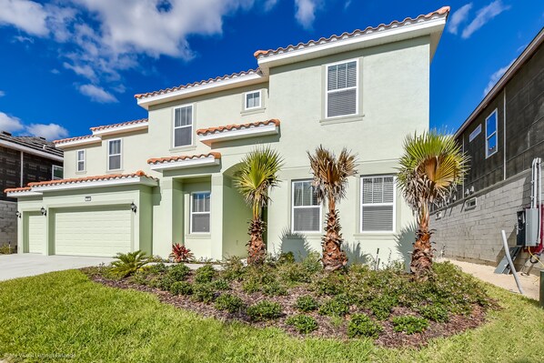 A two-story house with a light green exterior, a garage, and a well-maintained front yard with palm trees.