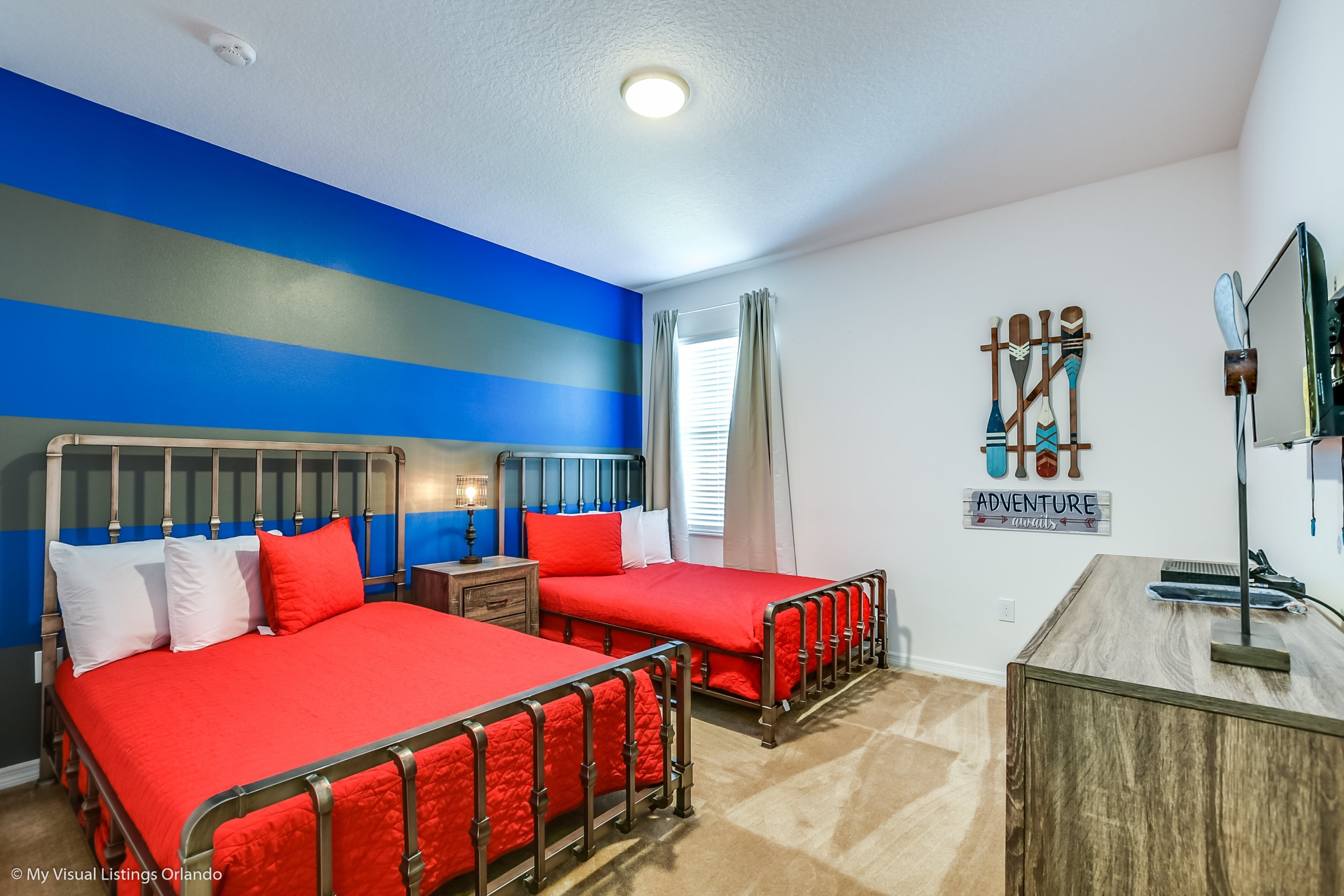 Quarto com duas camas, uma escrivaninha de madeira e uma parede com o letreiro 'Adventure Awaits'.