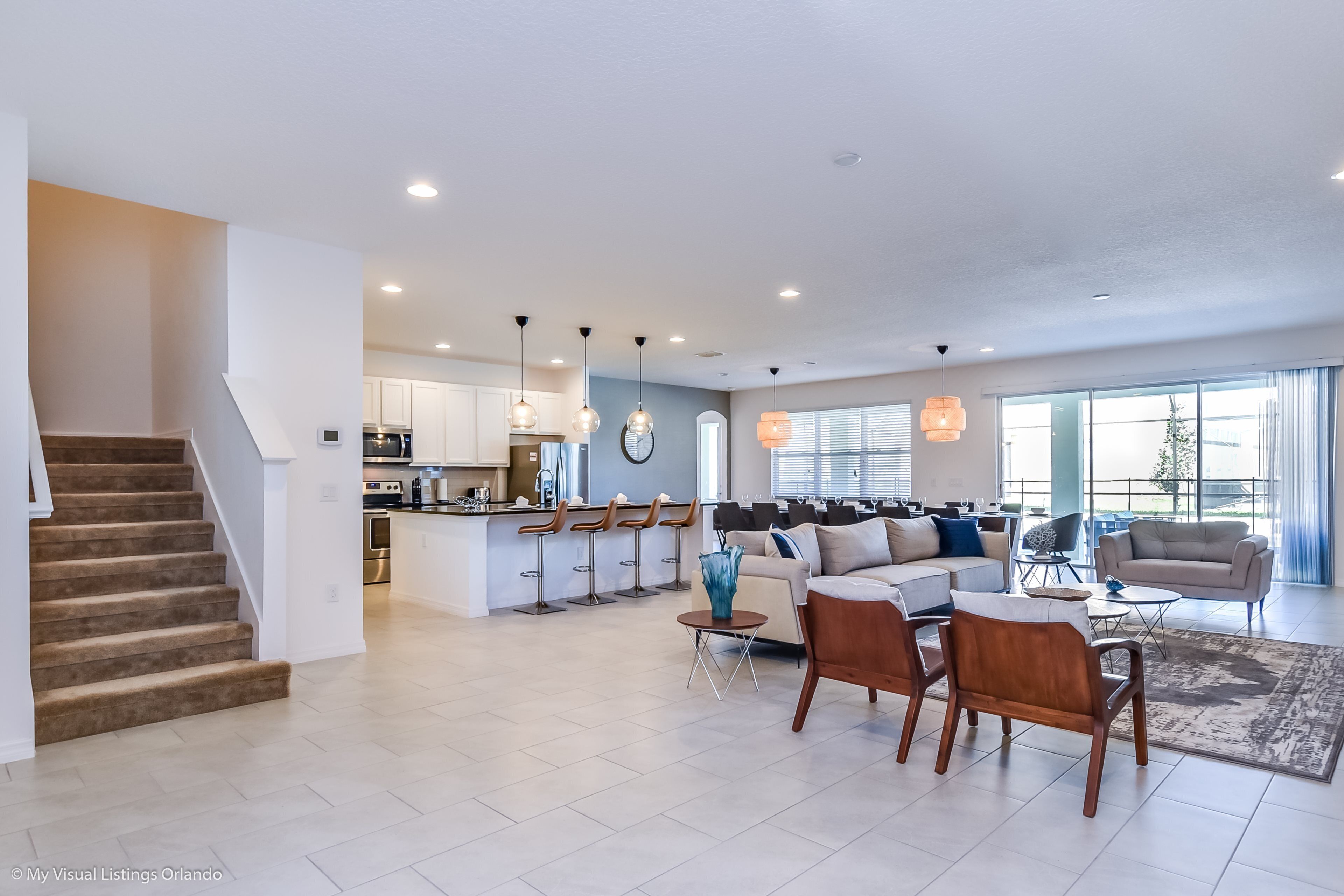 A spacious living area with a sofa, chairs, and a coffee table. A kitchen with modern appliances and bar stools is visible in the background.