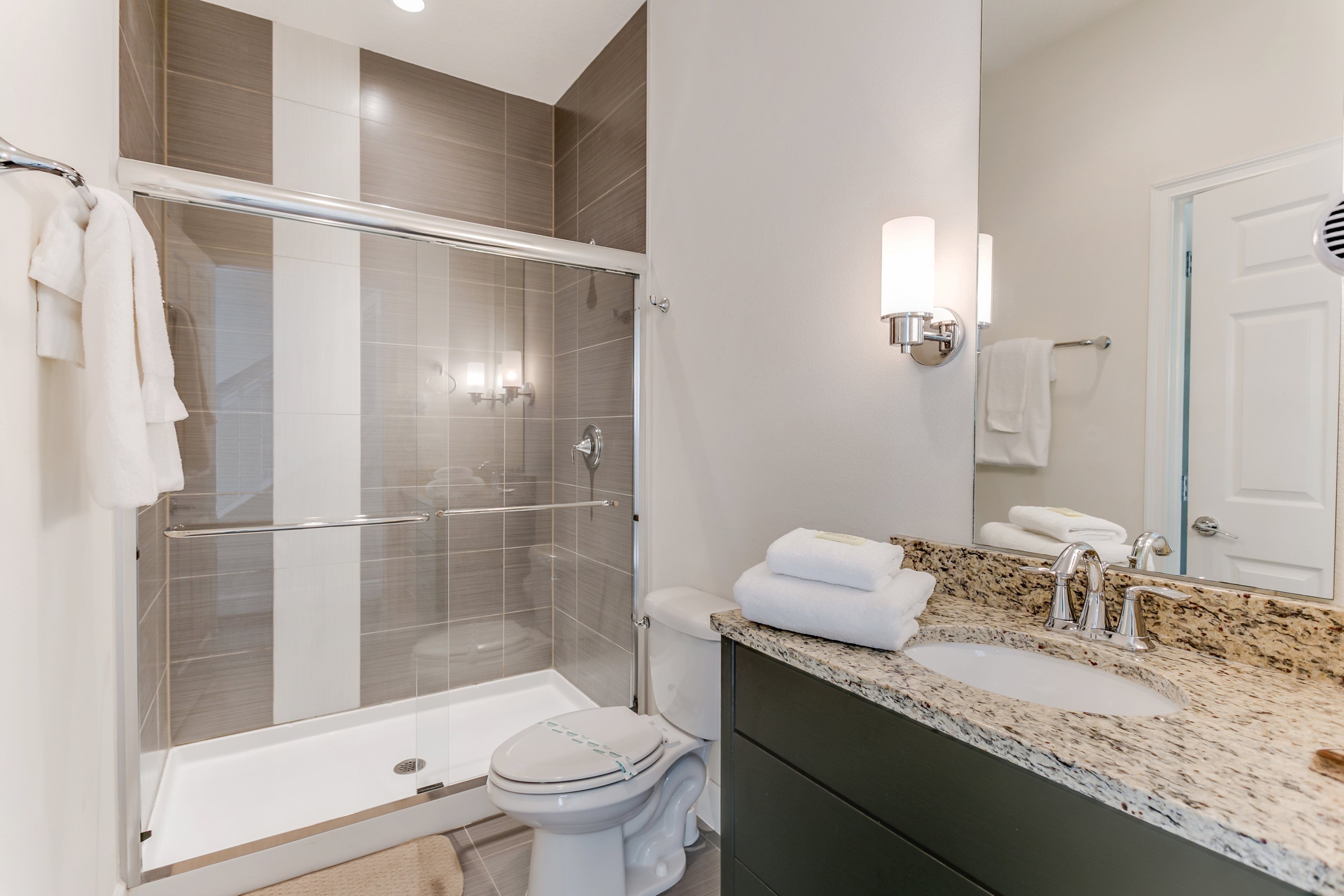 A modern bathroom with a glass-enclosed shower, a white toilet, a granite countertop sink, and a large mirror.
