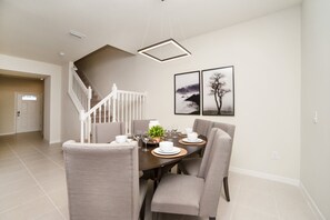 A dining area with a round table set for four, surrounded by chairs. A staircase with white railings leads to another room. Two framed pictures are on the wall.