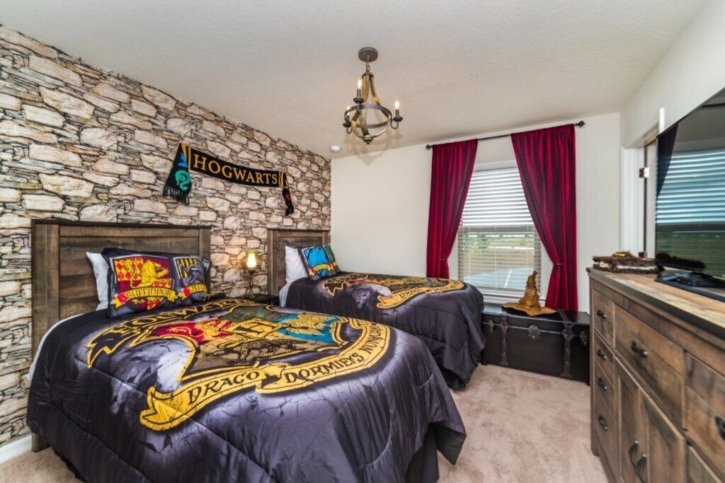 A room with a stone wall, two beds with Hogwarts-themed bedding, a chandelier, and a window with red curtains.