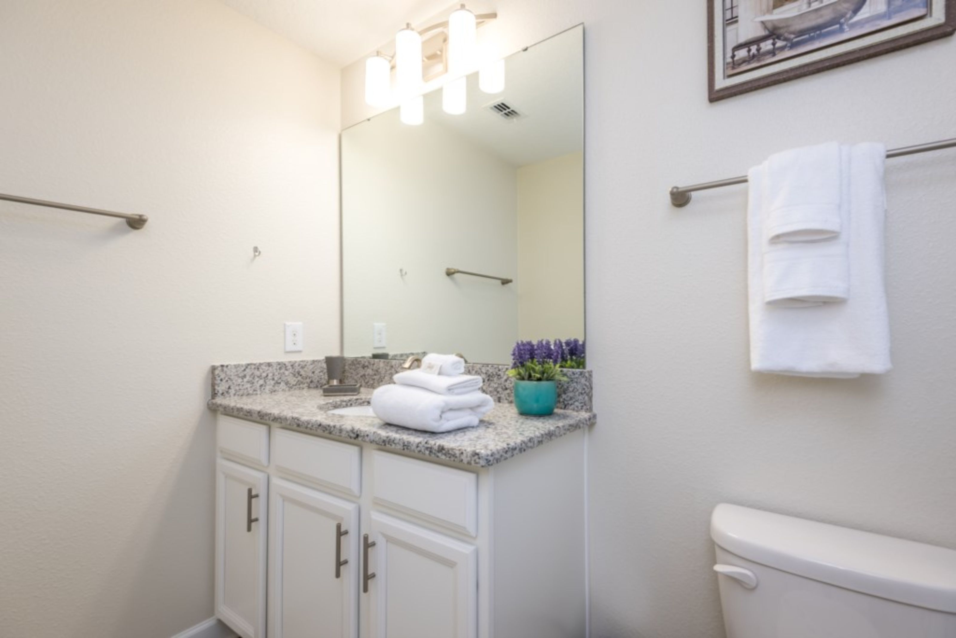 A bathroom with a granite countertop, white cabinets, a large mirror, a towel rack, and a toilet.