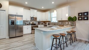 A modern kitchen with white cabinets, a stainless steel refrigerator, granite countertops, and a central island with bar stools. - 6 Bedroom 5 Bathroom Vacation Home (Davenport)
