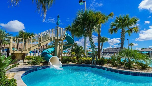 Una piscina con tobogán de agua, palmeras y un cielo azul despejado.
