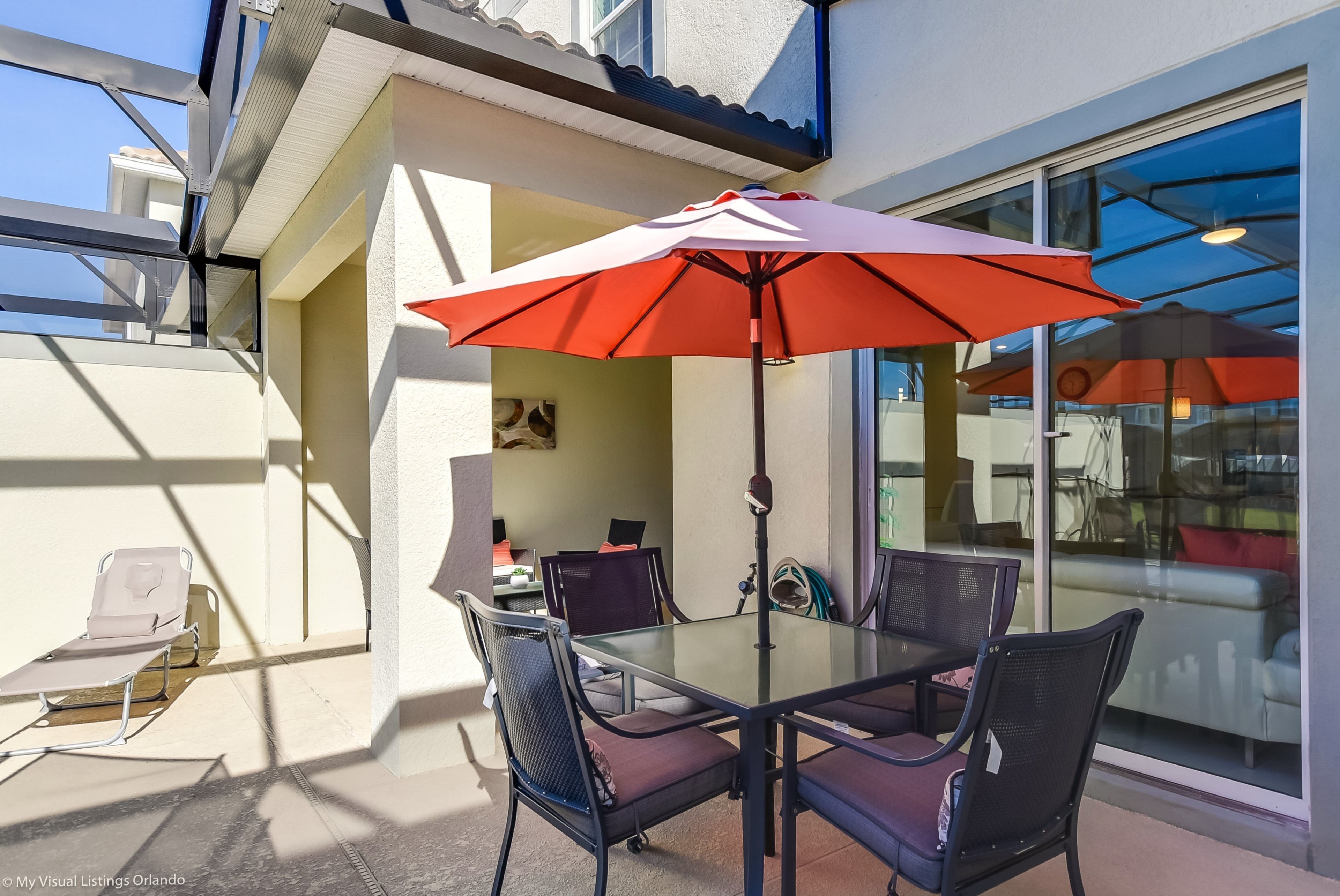 A glass-top table with four chairs and a red umbrella in a sunlit outdoor area with a view of a building.