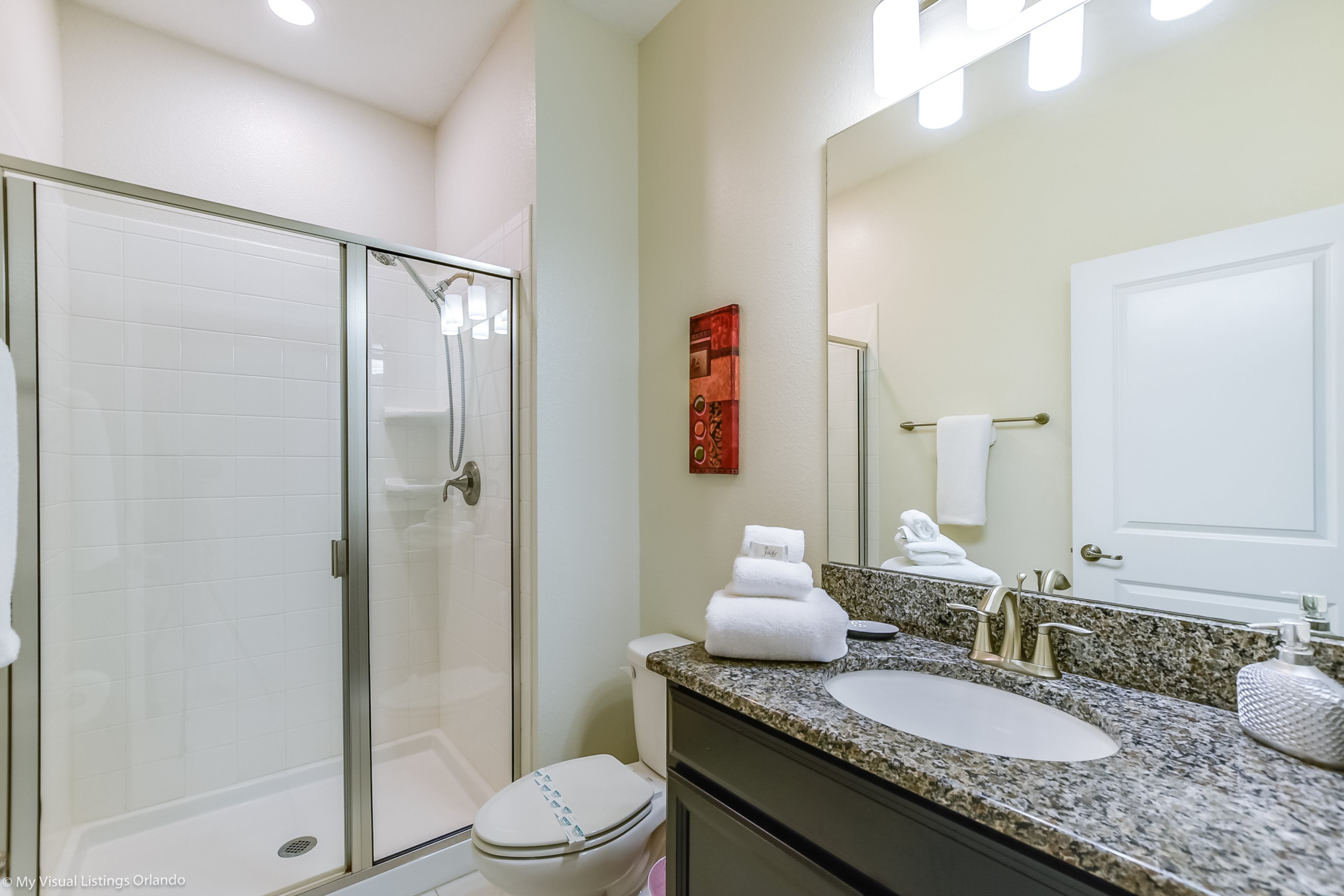 Une salle de bain avec une douche à parois de verre, des toilettes blanches, un comptoir en granit, un lavabo et un miroir.