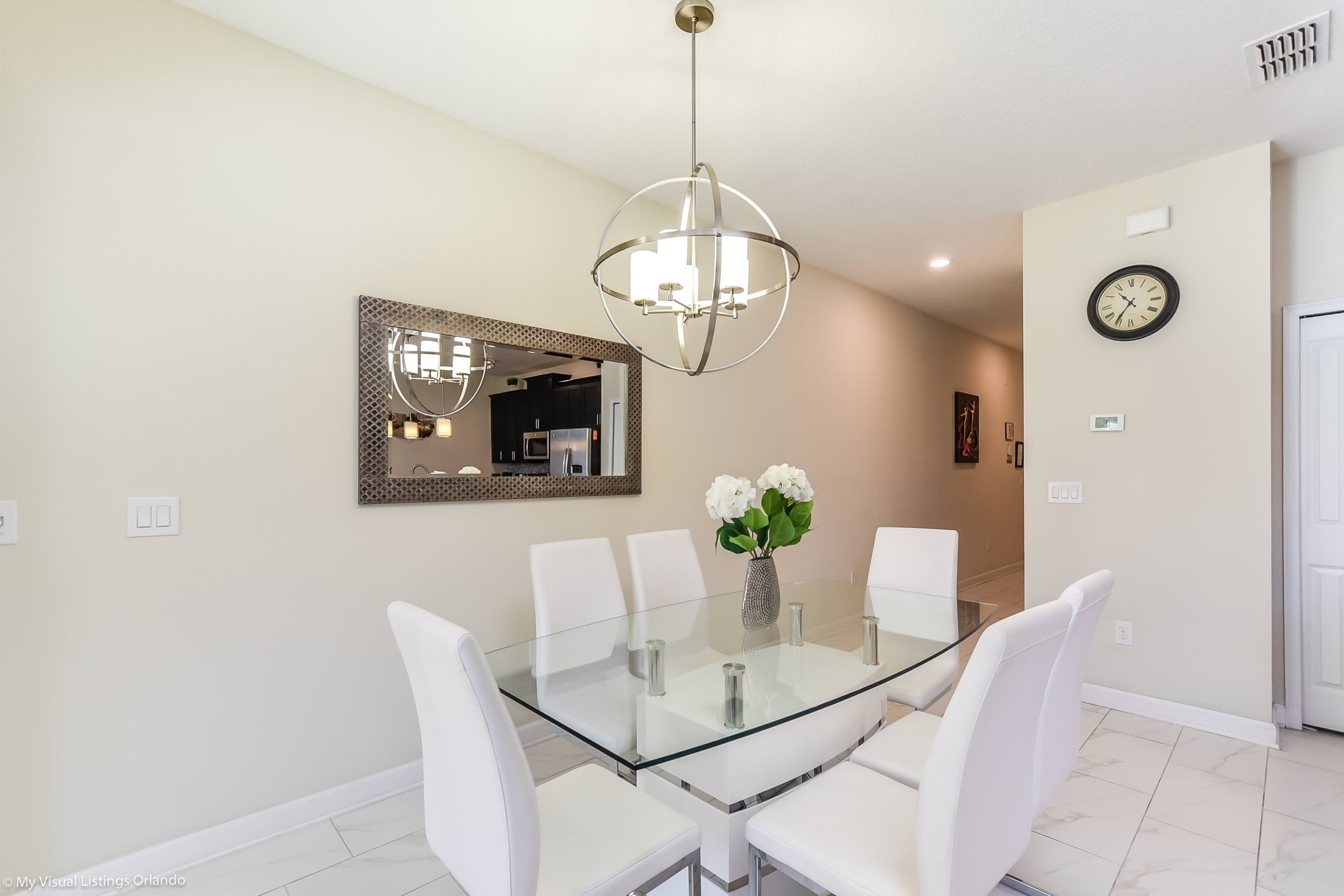 A dining area with a glass table surrounded by white chairs, a chandelier, a mirror, and a clock on the wall.