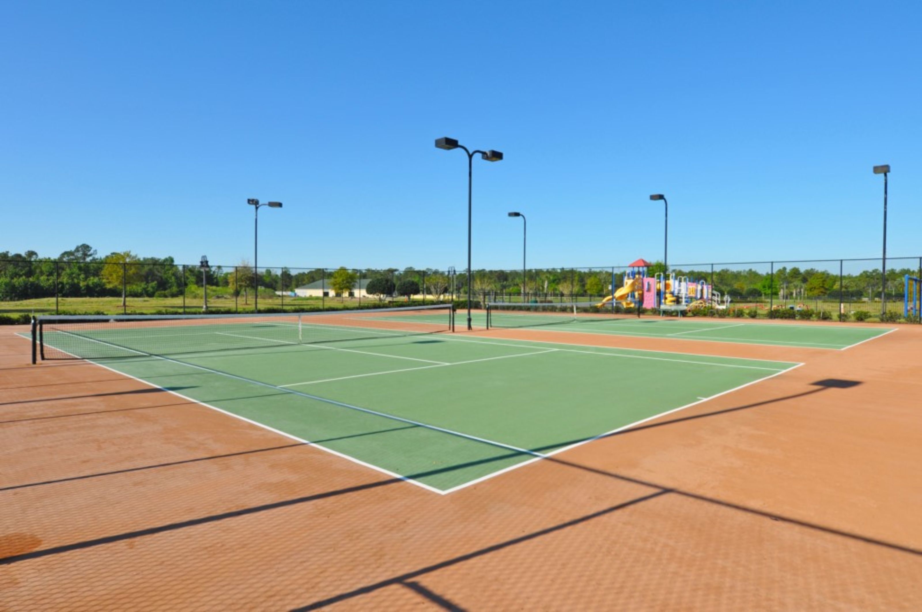 Una cancha de tenis con una red, rodeada por una cerca, y un parque infantil al fondo.