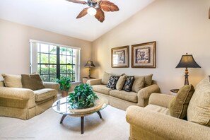 A living room with two beige sofas, a glass coffee table, a ceiling fan, and two framed pictures on the wall.