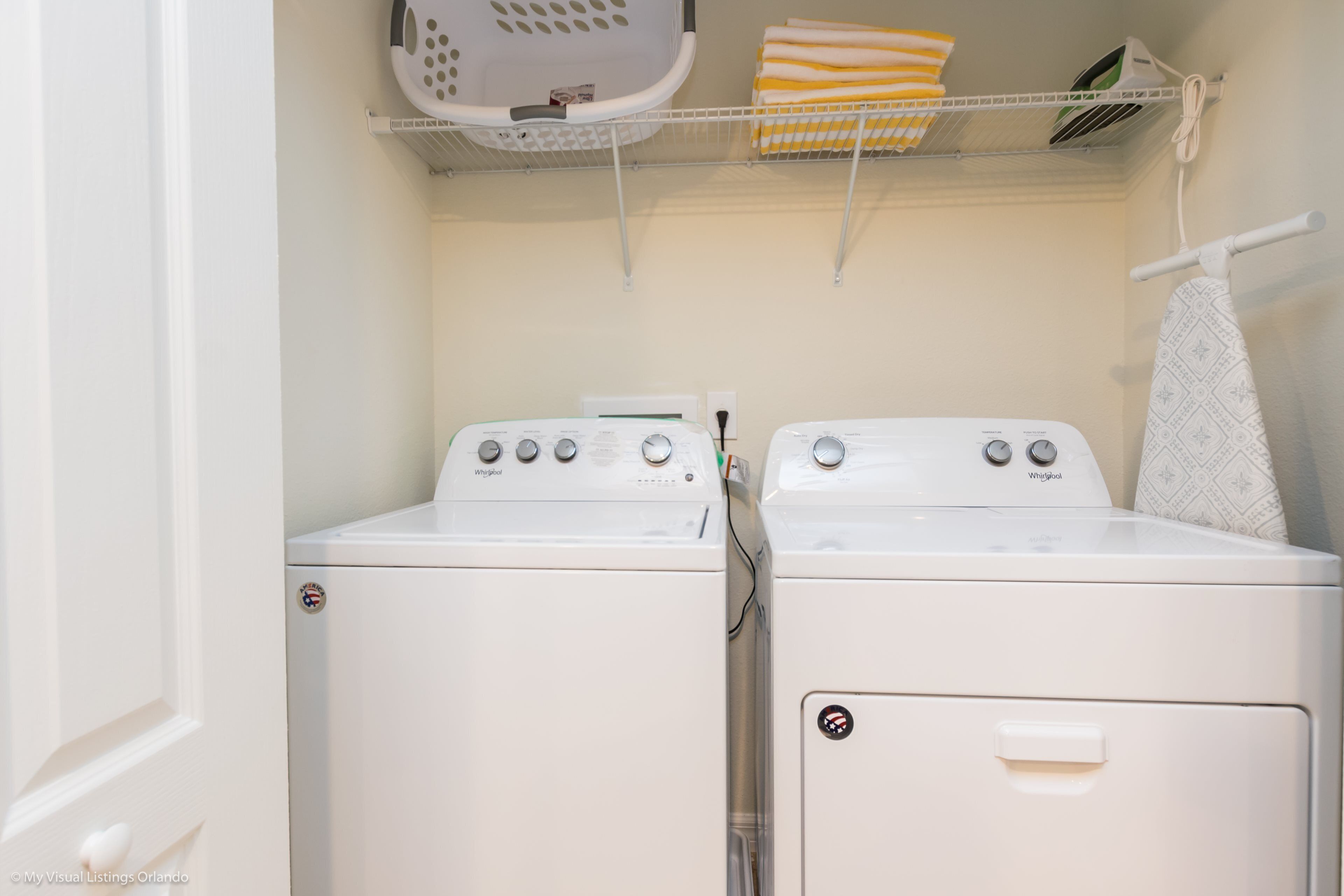 A laundry room with two white Whirlpool washers and dryers, a white basket, and a shelf with folded towels.