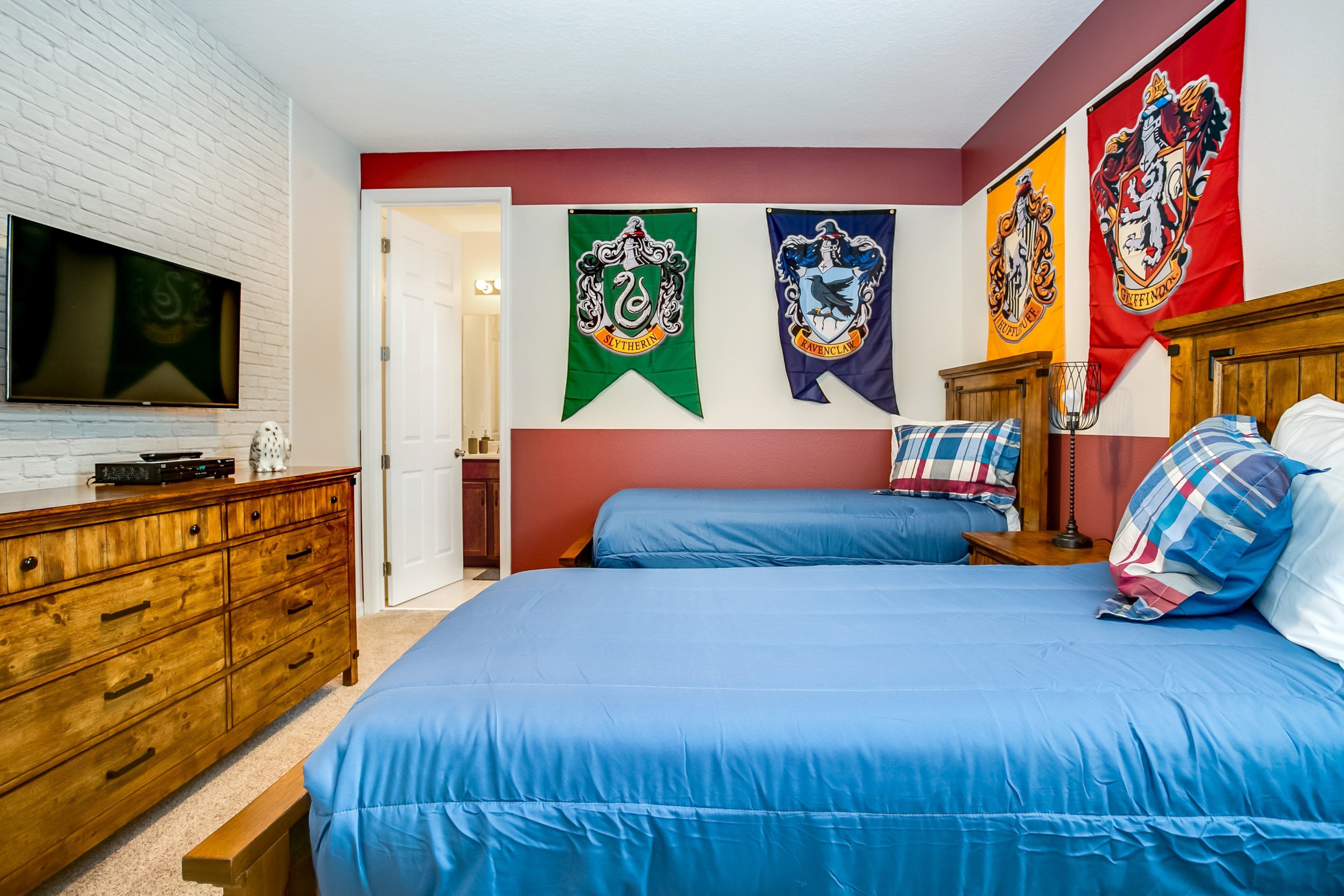 Un dormitorio con un aparador de madera, un televisor de pantalla plana, dos camas con ropa de cama azul y tres banderas en la pared.