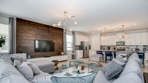 A modern living room with a large sectional sofa, a glass coffee table, and a flat-screen TV mounted on a wooden wall. - 9 BD 6.5 BH With Pool, Spa and GR (Kissimmee)