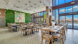 A spacious dining area with tables and chairs, a green wall with artwork, and large windows offering a view of buildings and a body of water. - Magic Village Resort 4 Bdrm Townhome (Kissimmee)