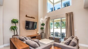 A spacious living room with a large flat-screen TV, grey sofas, a wooden coffee table, and potted plants. - Magic Village Resort 4 Bdrm Townhome (Kissimmee)